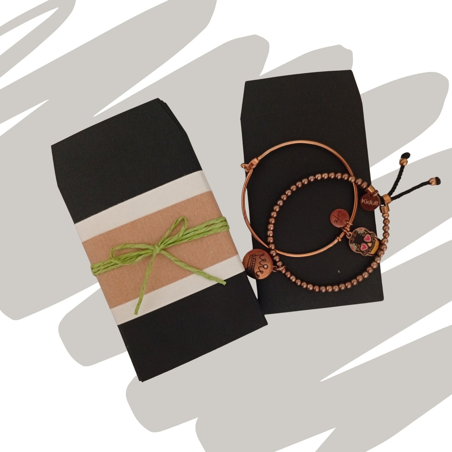 Due sacchetti neri posizionati su uno sfondo con strisce decorative beige e marroni. Un sacchetto è chiuso con una fascetta beige e un cordino verde con fiocco; accanto, due braccialetti artigianali in rame e perline sono appoggiati sul secondo sacchetto.