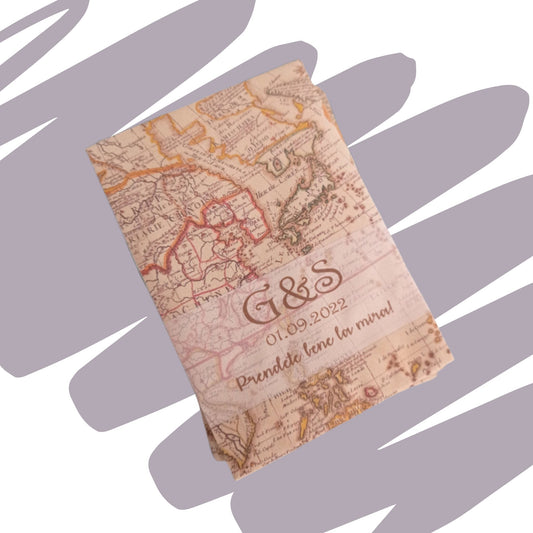 Sacchetto portaconfetti o bustina regalo in carta stampata con la grafica di una mappa geografica antica in stile vintage. Personalizzato con le iniziali "G&S", la data "01.09.2022" e la frase ironica "Prendete bene la mira!" stampate in color marrone seppia, posizionato su uno sfondo grigio con pennellate.
