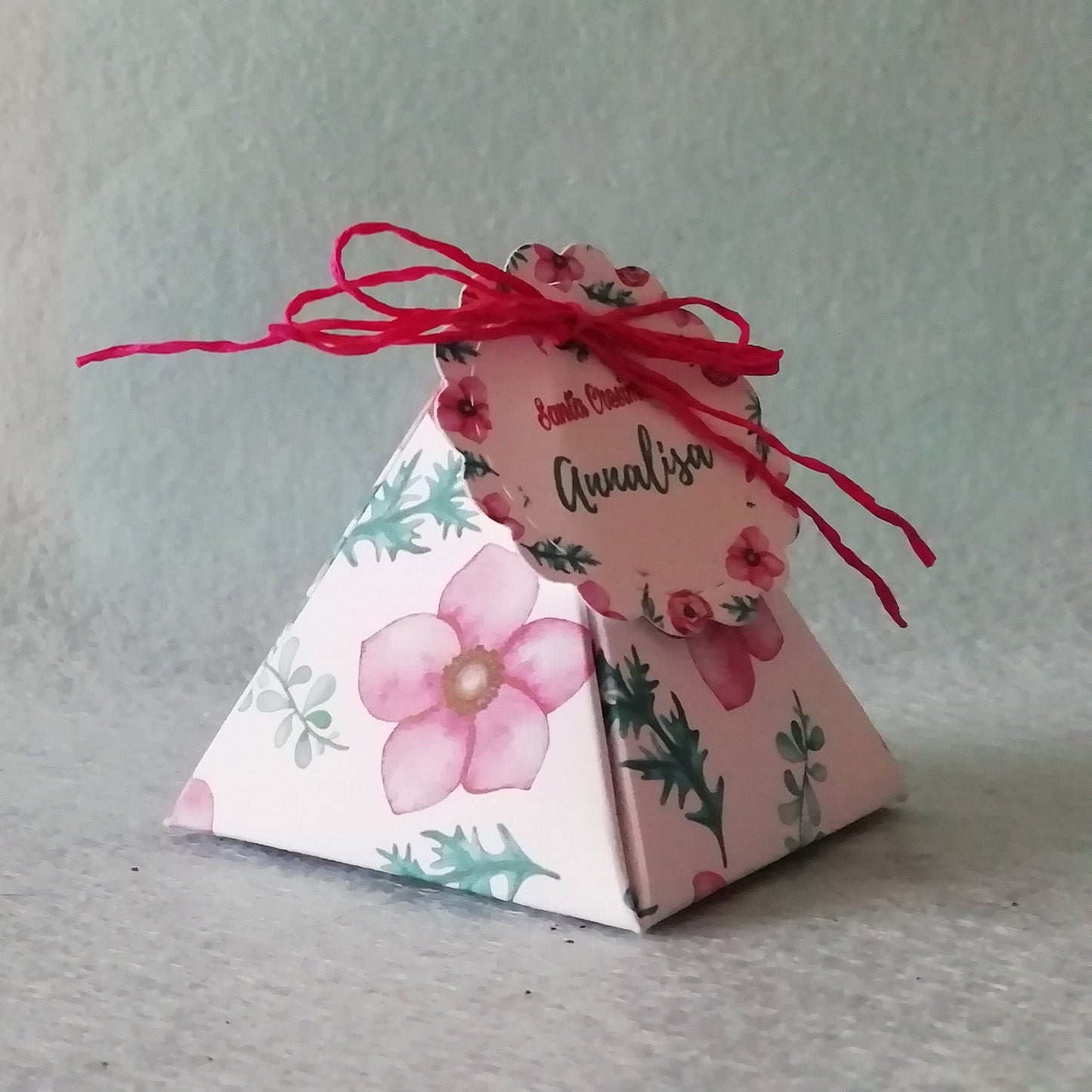 una scatolina portaconfetti con grandi fiori  rosaacceso e rametti di foglie con un tag rotondo personalizzato 